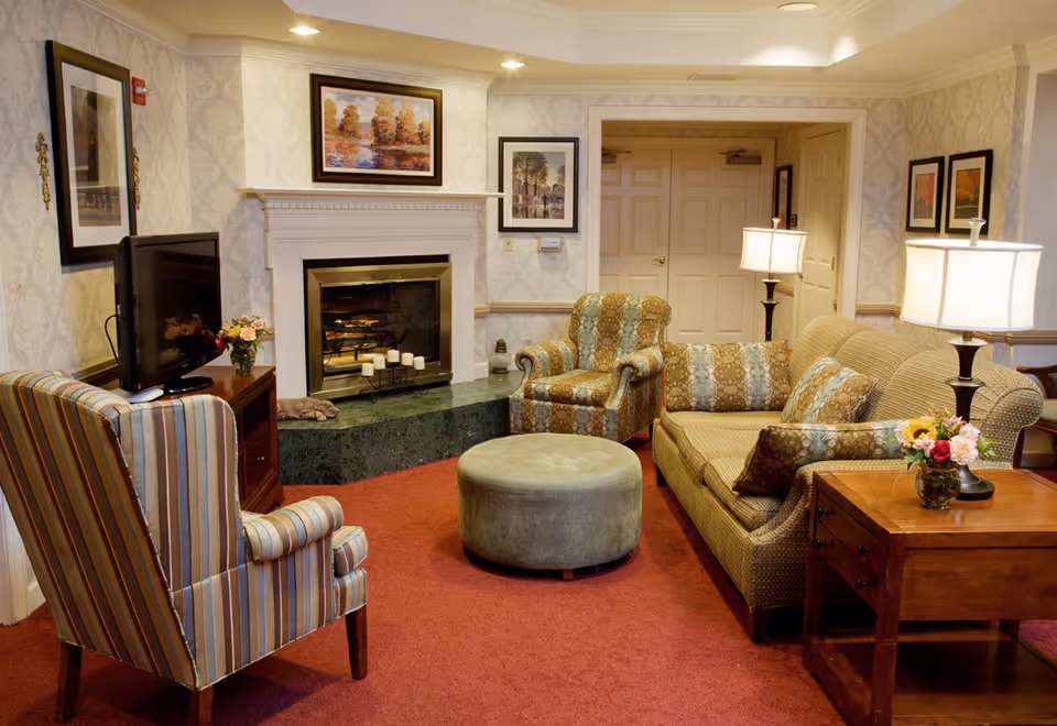 Cozy sitting area with a sofa, patterned armchairs, round ottoman, fireplace, TV and side tables with lamps and flowers.