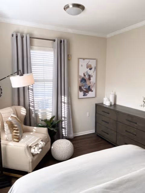 A cozy bedroom corner featuring a beige armchair with a pillow and a throw blanket, a round knitted ottoman, a floor lamp, and a window with gray curtains. A dark wooden dresser with decorative items and an abstract painting on the wall are also visible.