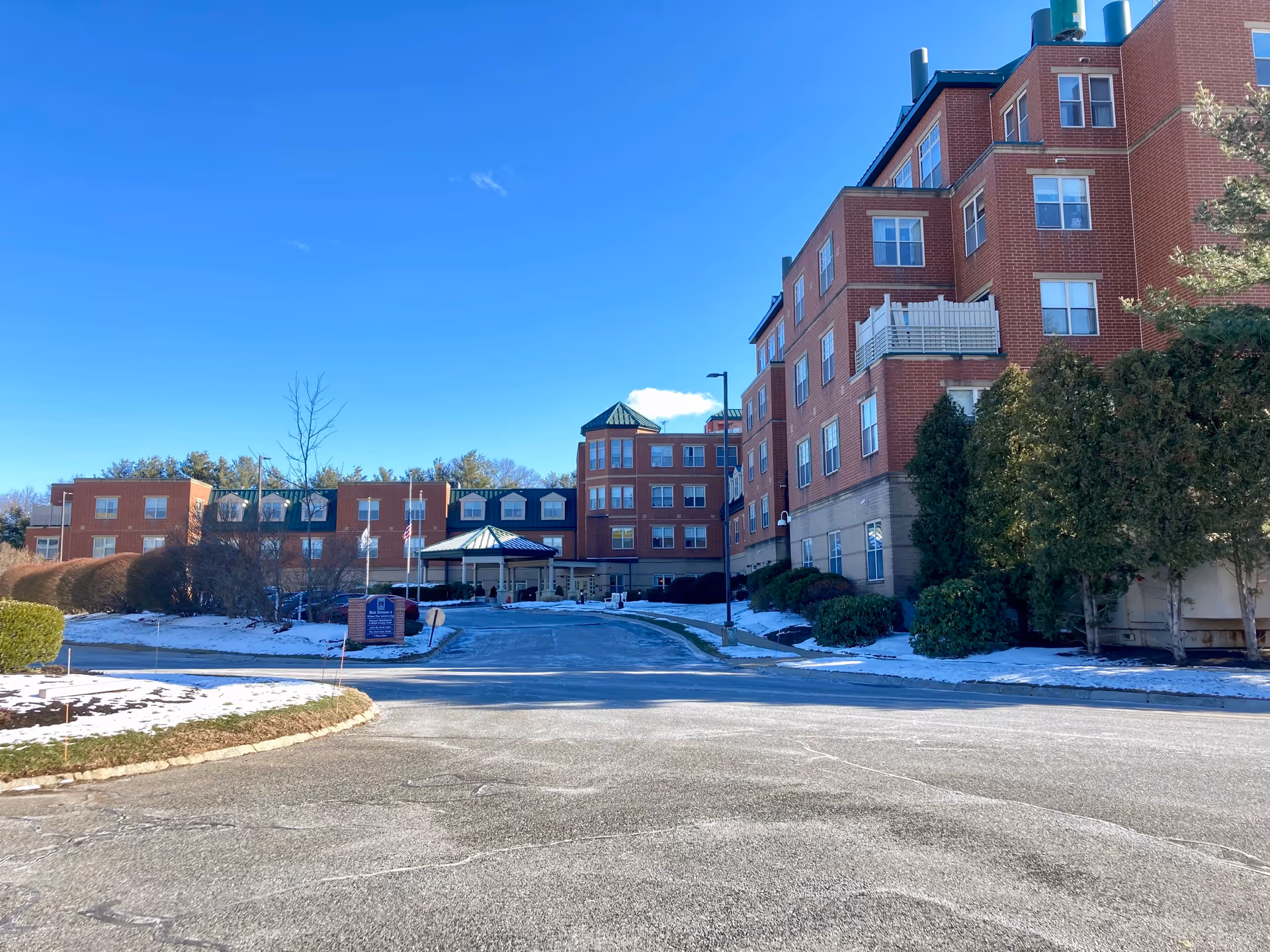 Exterior front view of a multi-story red brick assisted living building with an entrance canopy, circular driveway, and light snow on the ground.