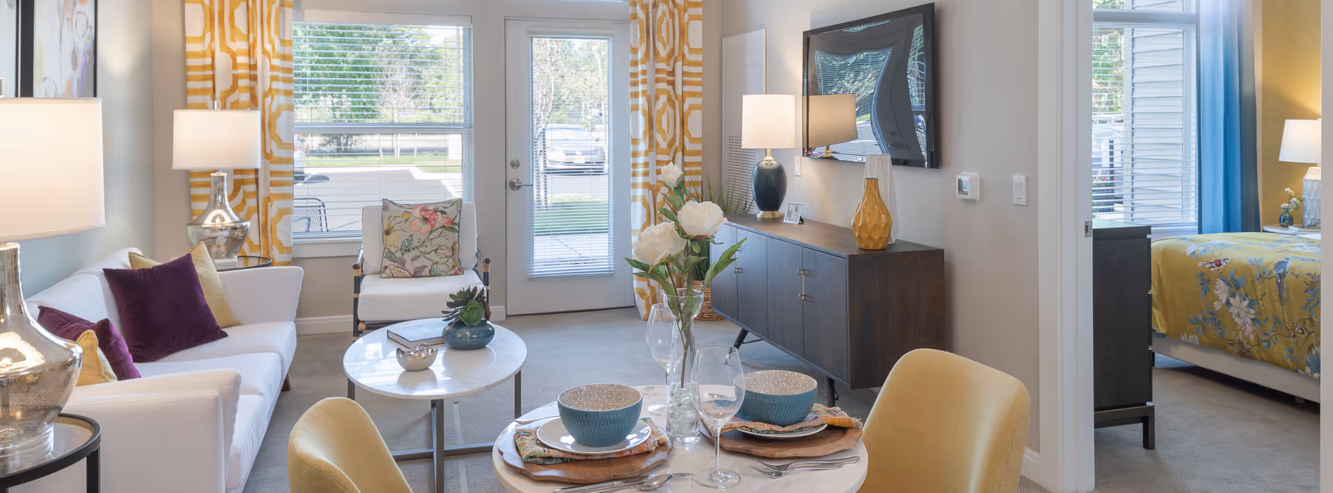 Bright and cozy senior living space featuring a white sofa with purple and yellow pillows, a small round coffee table with decorative items, a dining table set for two with bowls and glasses, a dark wood sideboard with lamps and a wall-mounted TV, large windows with yellow patterned curtains, and a glimpse into a bedroom with a yellow floral bedspread.