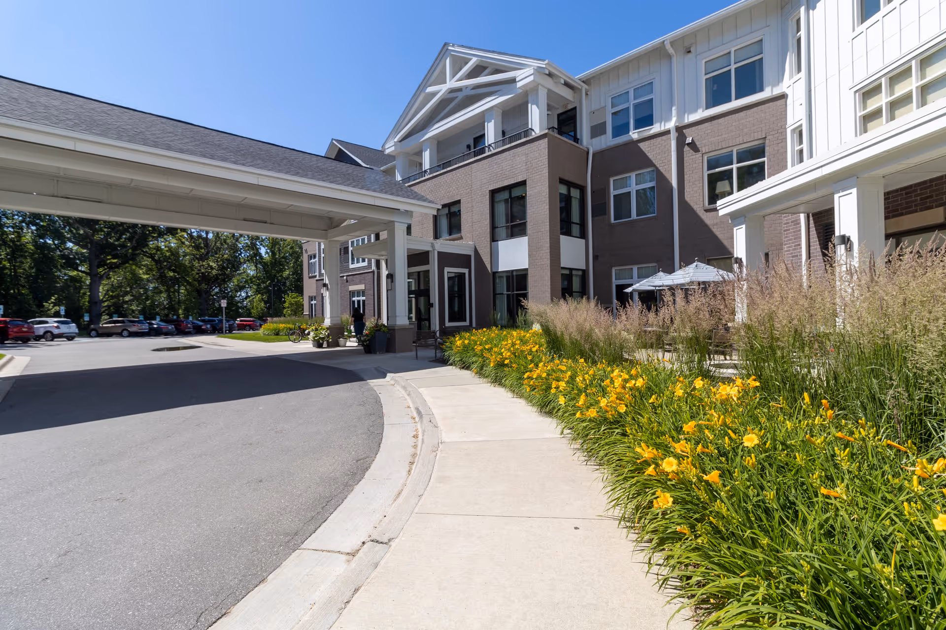Exterior view of StoryPoint Novi senior living facility showing the entrance with a covered drop-off area, a sidewalk lined with yellow flowers and ornamental grasses, and a parking lot with several cars. The building is three stories tall with large windows and a mix of brick and white siding.