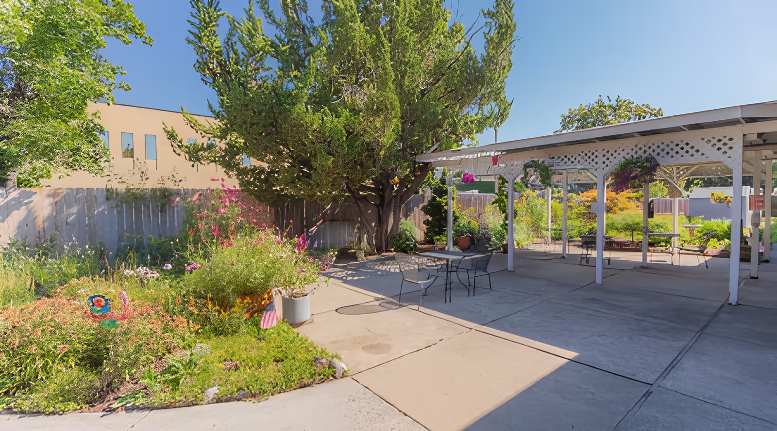 Outdoor patio area at Hearthstone Nursing & Rehabilitation Center with a concrete floor, metal table and chairs, surrounded by greenery including trees, bushes, and flowering plants. There is a white pergola structure providing partial shade.