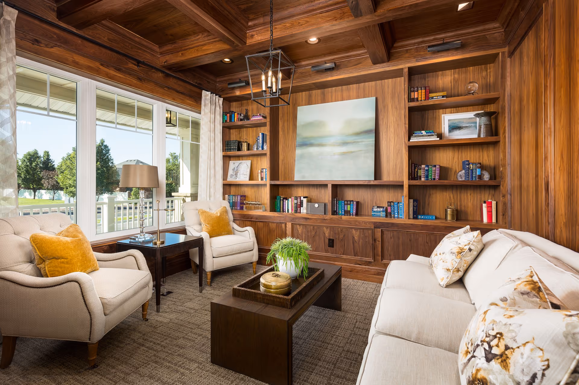 A cozy living room with wooden paneled walls and built-in bookshelves filled with books and decorative items. The room features a beige sofa with floral pillows, two beige armchairs with mustard yellow pillows, a dark wooden coffee table with a plant and decorative items, a side table with a lamp, and large windows with white curtains letting in natural light and offering a view of green trees outside.