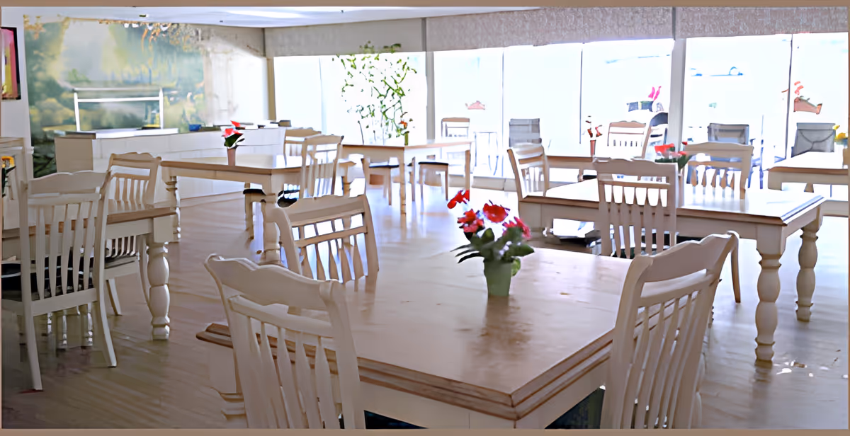 A bright dining room with multiple wooden tables and chairs arranged neatly. Each table has a small vase with red flowers. Large windows in the background allow natural light to fill the room, and there is a mural on one wall depicting a nature scene.
