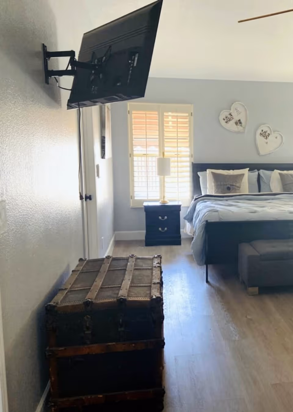 Bedroom with a wall-mounted TV, a wooden trunk at the foot, nightstand and lamp by a bed beneath shuttered windows.