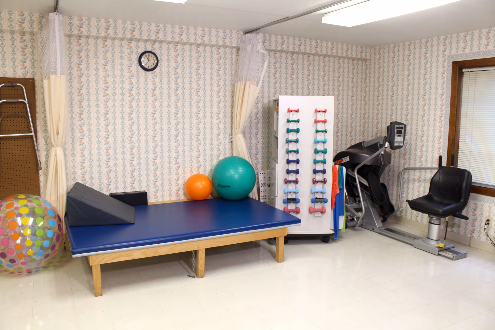 A physical therapy or exercise room with a blue padded therapy table, various exercise balls including a large colorful polka dot ball, a rack holding multiple pairs of small dumbbells, and a seated exercise machine. The room has floral patterned wallpaper, a clock on the wall, and a window with blinds.