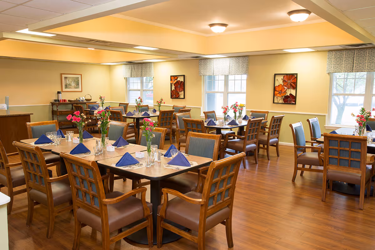 Communal dining room with wooden tables and chairs set with blue napkins and vases of flowers.