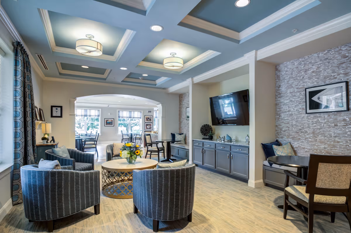 Bright communal lounge with curved armchairs around a central table, a wall-mounted TV above built-in cabinets, and a dining area visible through an archway.