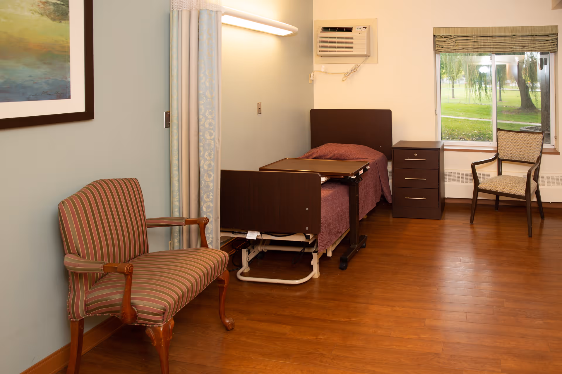 A simple bedroom in a senior living facility featuring a single hospital-style bed with a purple blanket, an overbed table, a wooden nightstand with drawers, a striped upholstered armchair, and a window with a view of green trees outside. The room has wooden flooring and light-colored walls with a framed painting and a wall-mounted air conditioning unit.