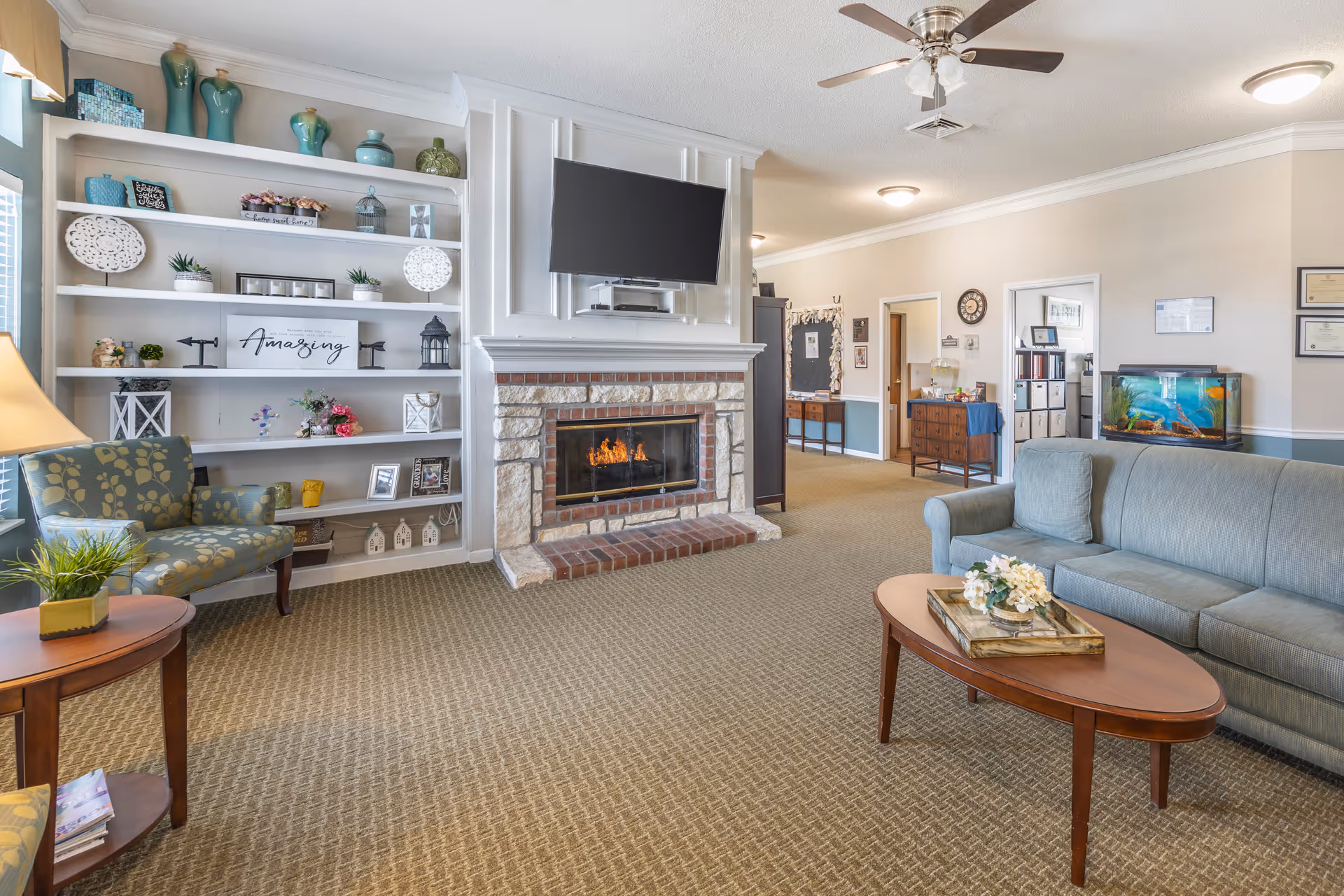 A cozy living room area in a senior living facility featuring a stone fireplace with a mounted flat-screen TV above it. To the left, there is a built-in white bookshelf decorated with vases, plants, and framed pictures. A patterned armchair and a wooden side table with a lamp and a plant are positioned near the bookshelf. On the right side, there is a gray sofa and a wooden coffee table with a floral arrangement. The room has beige walls, carpeted flooring, and a ceiling fan with lights. In the background, there is a hallway with framed certificates on the wall and a fish tank on a cabinet.