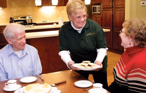 A staff member serves a plate of cookies to two seated elderly residents at a dining table in a kitchen/dining area.