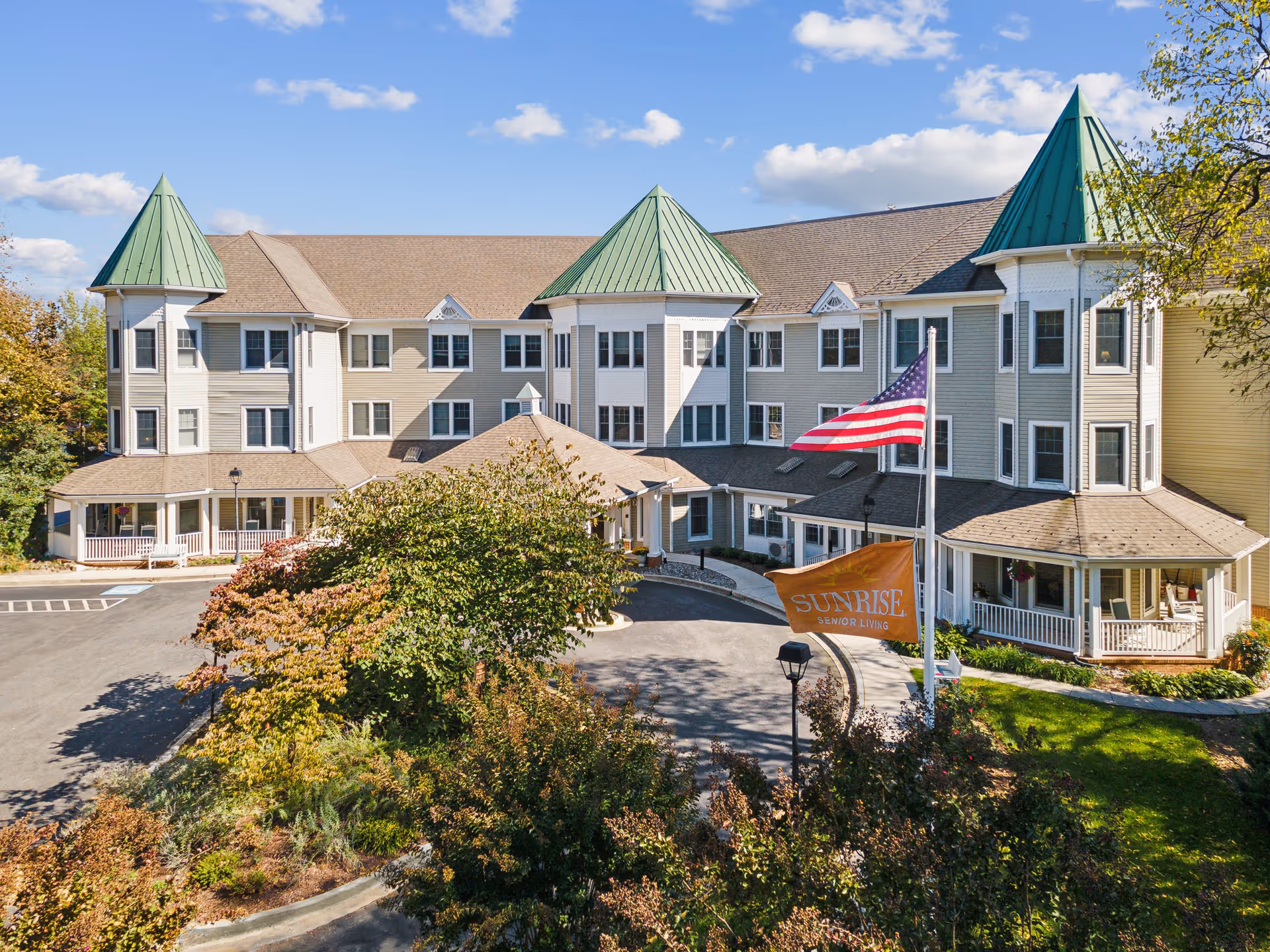 Front exterior of the Sunrise senior living building with green-roofed turrets, an American flag, and a circular entrance drive.