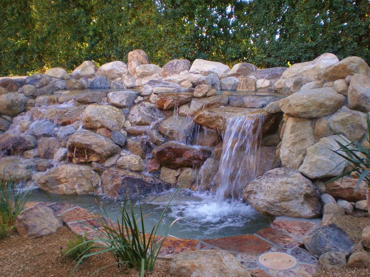 A small outdoor waterfall feature with water cascading over large natural rocks into a shallow pool, surrounded by greenery and plants.