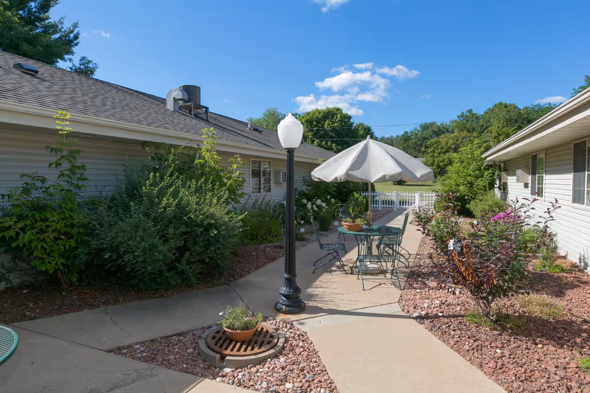 Courtyard patio between two single-story buildings with a table and umbrella, lamp post, and landscaping under a blue sky.