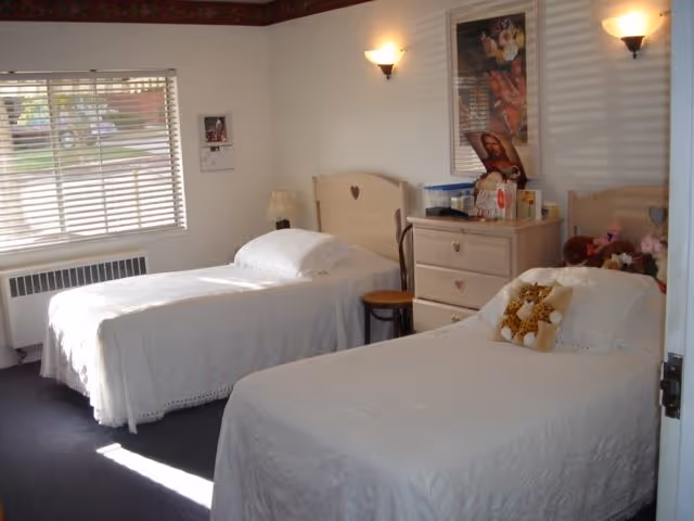 A bedroom with two single beds covered in white bedspreads. Each bed has a wooden headboard with a heart cutout. There is a small wooden dresser between the beds with various items on top, including a framed picture of Jesus. A window with blinds lets in natural light, and the walls are white with two wall-mounted lamps.