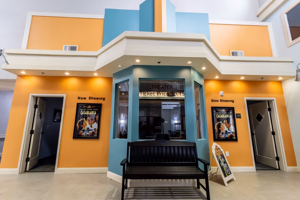 Interior view of a small theater entrance area with orange and blue walls. There are two open doors on either side, each with a movie poster for Casablanca and a sign above saying 'Now Showing'. In the center is a ticket booth with a sign indicating ticket price of $0.15 and a black bench in front of it. A small chalkboard sign is placed near the right door.
