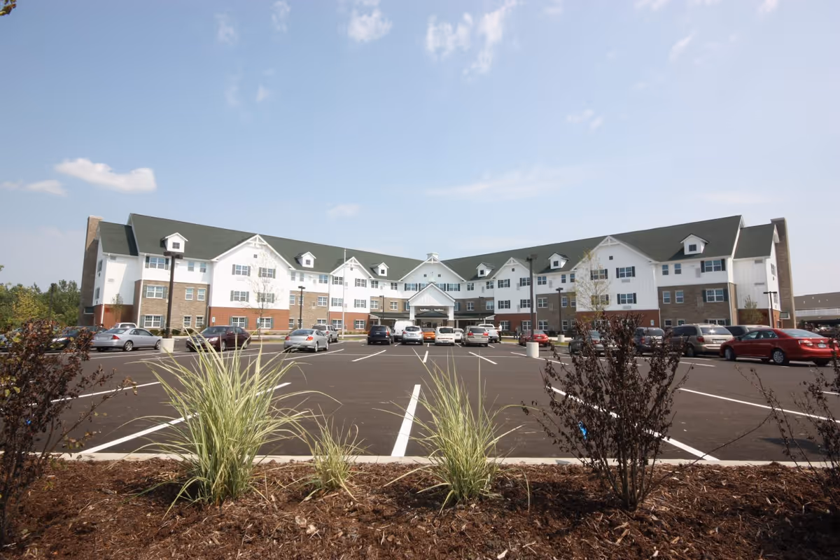 Exterior view of a large three-story senior living facility building with a green roof and white and brick facade, surrounded by a parking lot with several parked cars and landscaped plants in the foreground under a partly cloudy sky.
