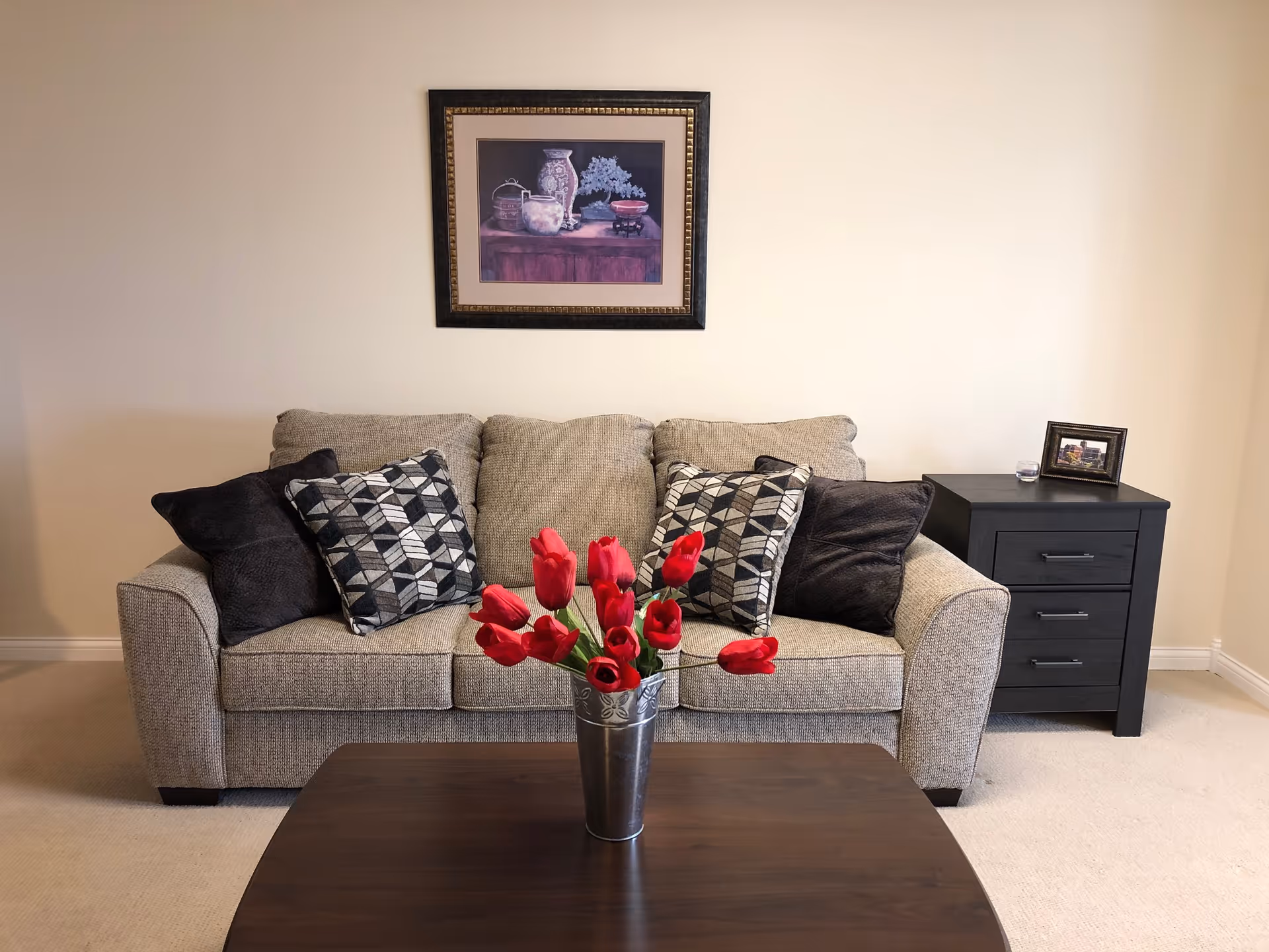 Beige couch with patterned throw pillows and a vase of red tulips on a coffee table in a simple living room with a framed picture and a black side table.
