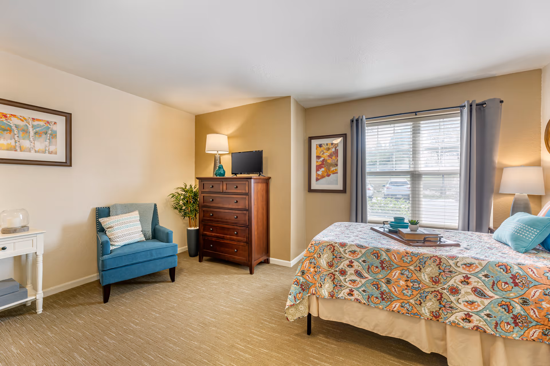 A cozy bedroom with a bed covered in a colorful patterned quilt, a wooden dresser with a small TV and lamp on top, a blue armchair with a pillow, a small white side table with a candle, and two framed artworks on the walls. A window with gray curtains lets in natural light.