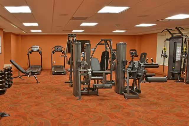 Fitness room with multiple weight machines, treadmills, dumbbells, and an orange patterned carpet.