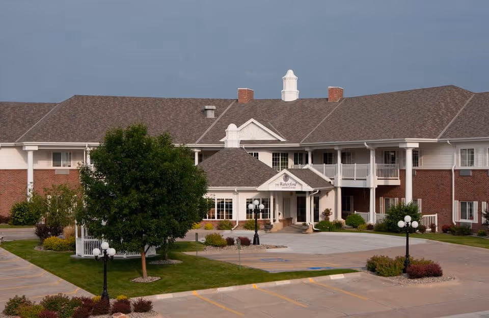 Exterior view of Waterford at Williamsburg senior living facility showing a large two-story building with a brick and white siding facade, a covered entrance, landscaped greenery, trees, and a parking lot in front.