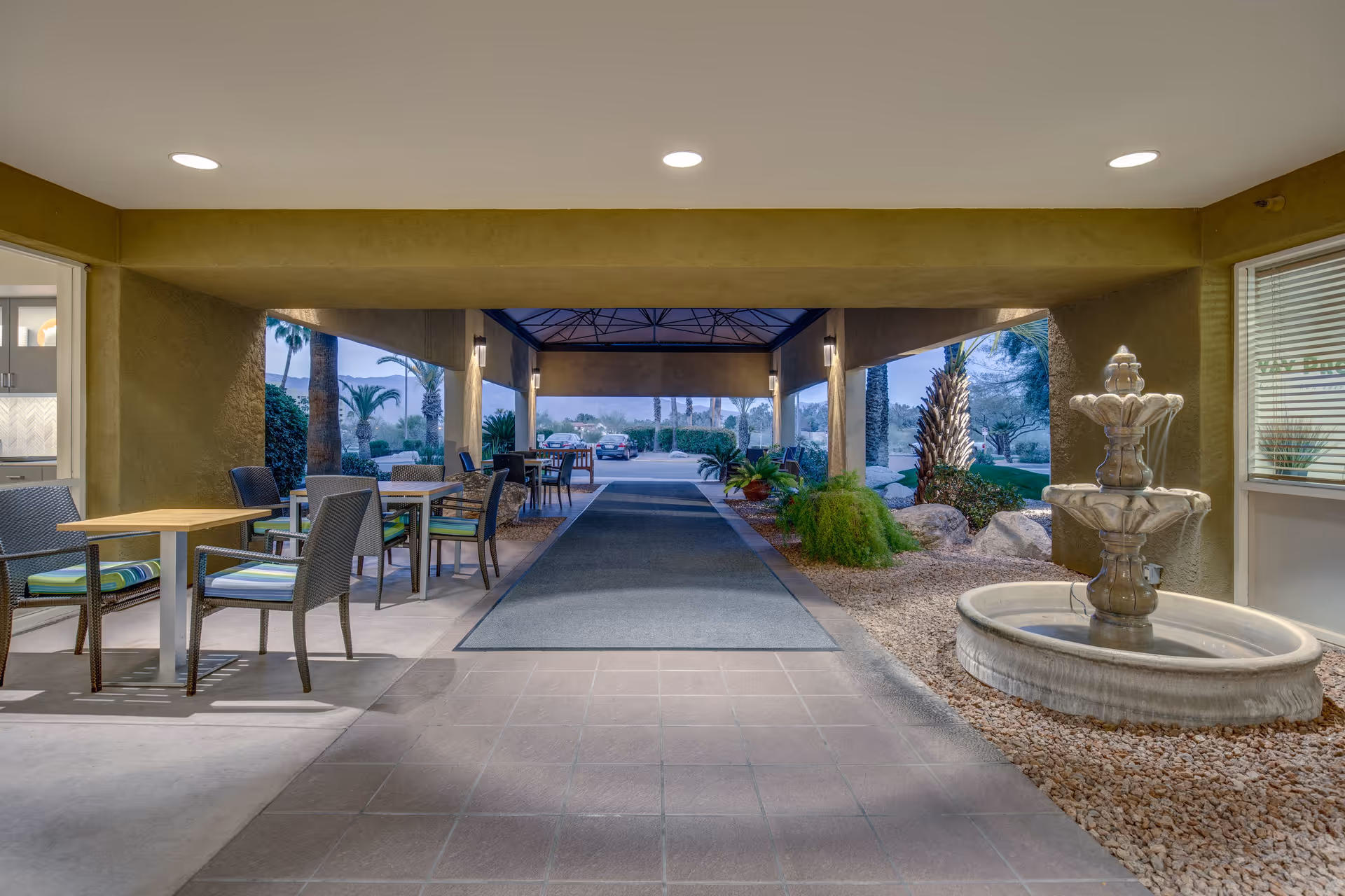 Covered outdoor seating area with tables and chairs on the left side, a stone water fountain on the right, and a walkway leading to a parking area with palm trees and landscaping visible in the background.