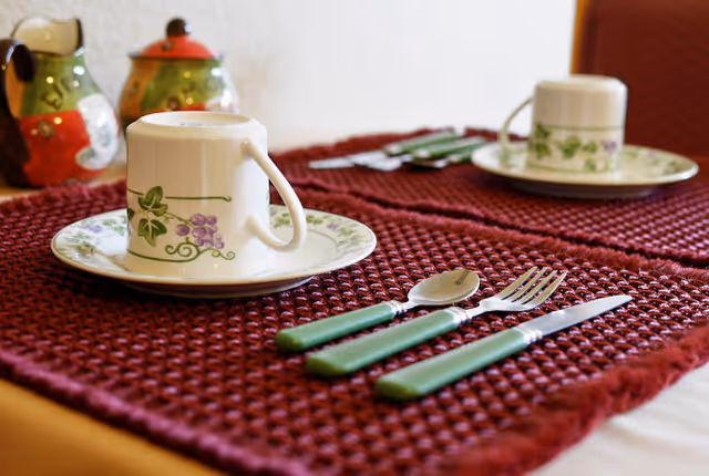 Close-up of a dining table setting with a white cup and saucer decorated with green leaves and purple grapes, placed on a textured dark red placemat. Green-handled fork, knife, and spoon are arranged beside the cup. In the background, there are two small decorative containers shaped like owls.