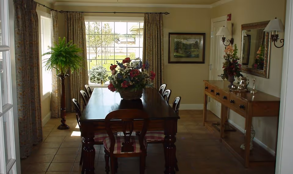 Dining room with a wooden table and chairs, a floral centerpiece, sideboard, mirror and a large window with curtains.