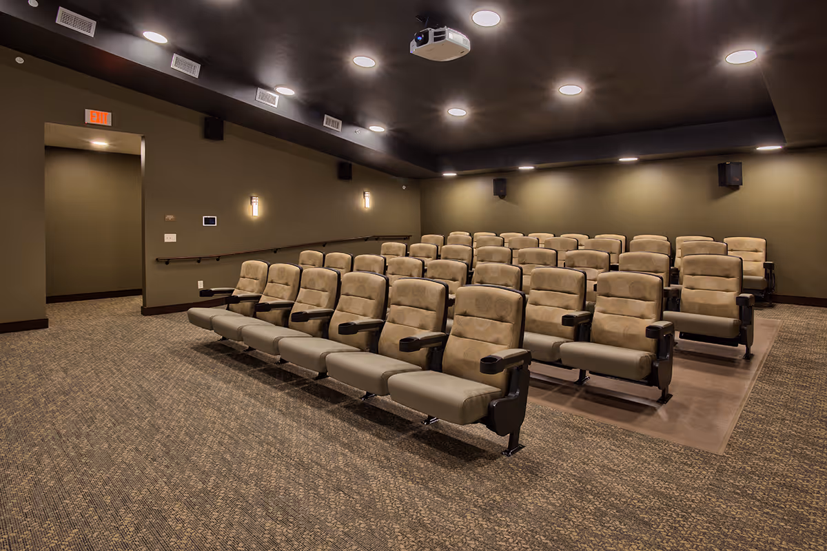 A small theater room with multiple rows of beige cushioned seats with armrests, a dark ceiling with recessed lighting, a projector mounted on the ceiling, and olive green walls.