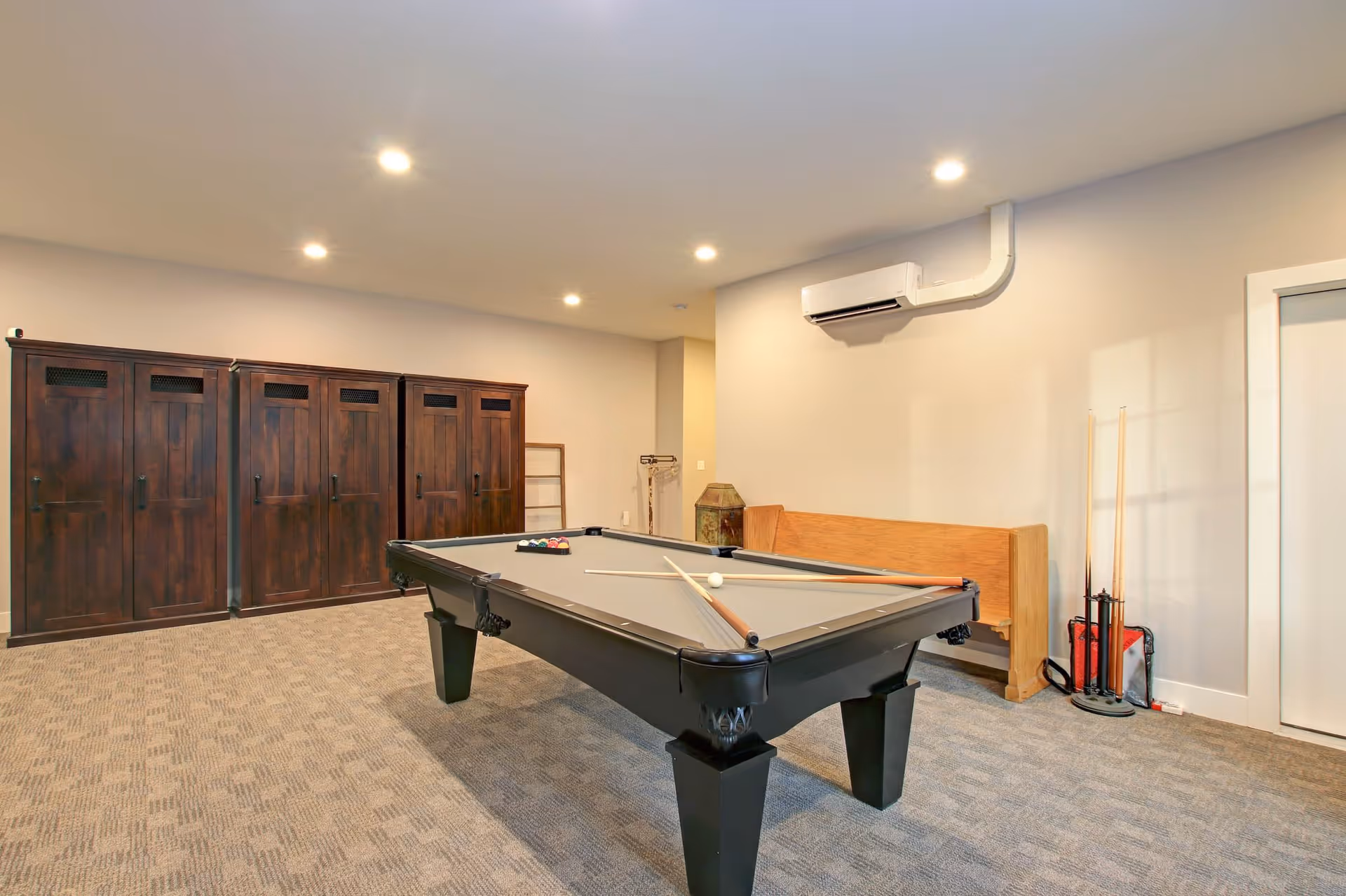 A recreational room with a pool table in the center, pool cues resting on the table, a wooden bench against the wall, and several dark wooden lockers along the back wall. The room has beige walls, carpeted floor, and recessed ceiling lights.