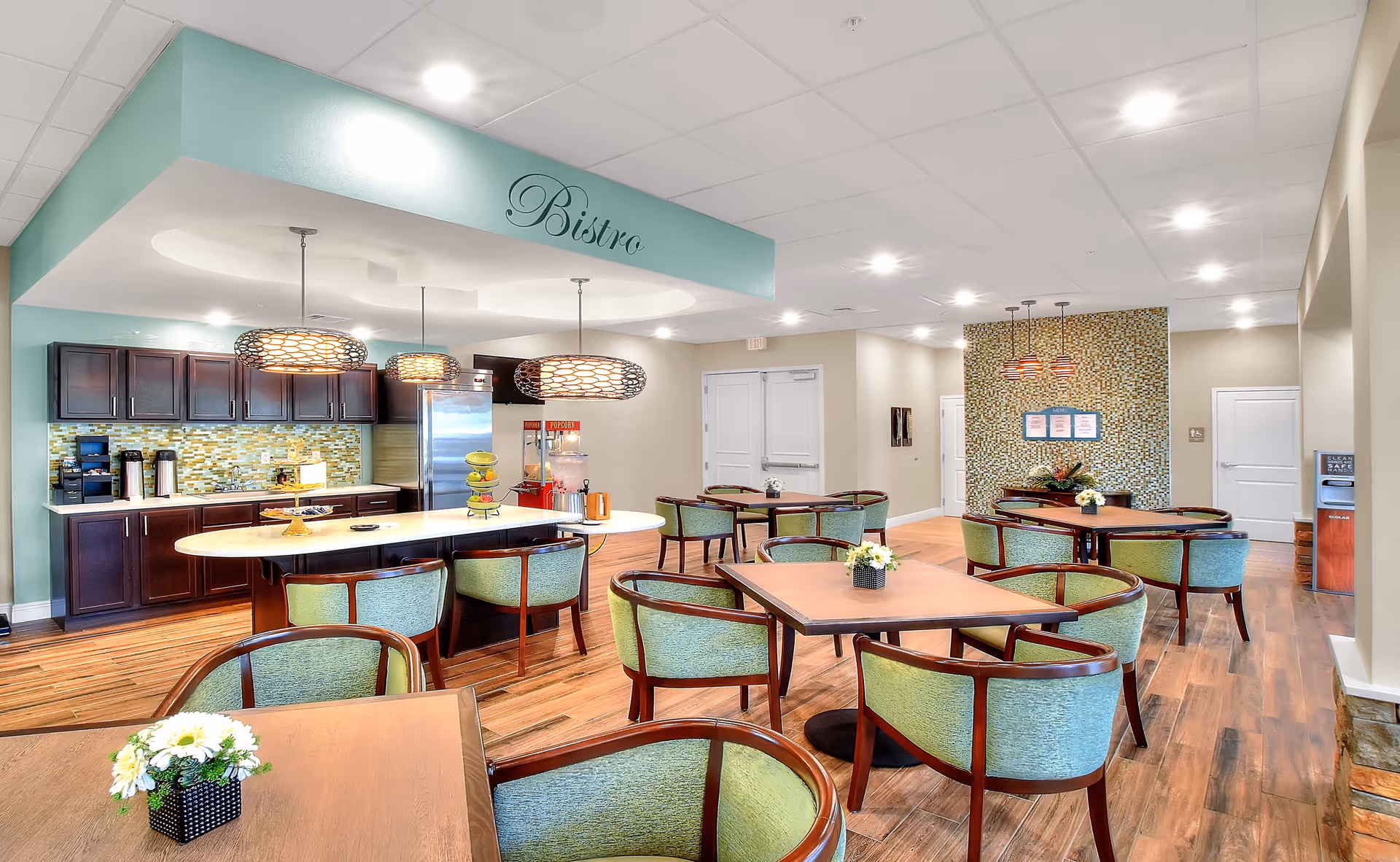 A bright and modern bistro area in a senior living facility featuring wooden tables with green cushioned chairs, a kitchen counter with dark cabinets, a stainless steel refrigerator, a popcorn machine, and decorative pendant lights hanging from the ceiling. The space has wood flooring and a mosaic tiled accent wall in the background.