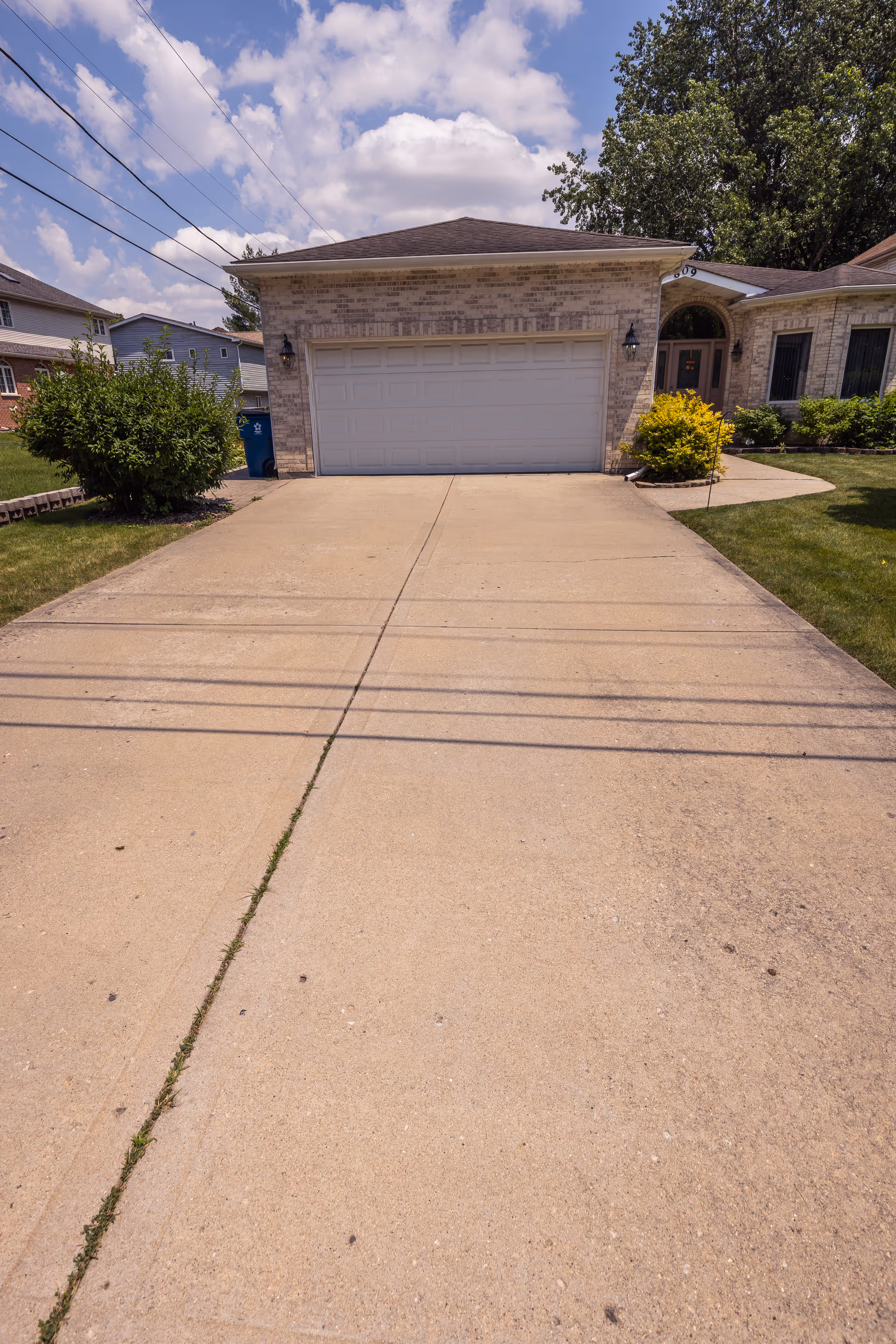 A concrete driveway leading up to a two-car garage attached to a single-story brick house. The house has a dark shingled roof, two outdoor wall lanterns flanking the garage door, and a front entrance with an arched window above the door. There are green bushes and a small tree on either side of the driveway, and a blue recycling bin is visible near the left side of the garage. The sky is partly cloudy with blue patches.