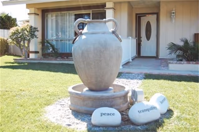 Outdoor garden area in front of a building entrance with a large decorative urn fountain surrounded by three white stones inscribed with the words 'peace', 'tranquility', and 'harmony'.