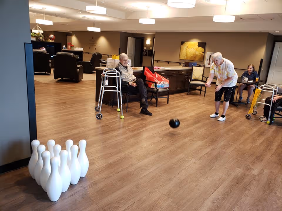 An elderly woman is playing indoor bowling in a spacious room with wooden flooring. She is rolling a black bowling ball towards white bowling pins set up on the floor. Several elderly people are seated around the room, some with walkers nearby, watching the game. The room is well-lit with ceiling lights and has comfortable chairs and a cabinet with a yellow flower painting on the wall.