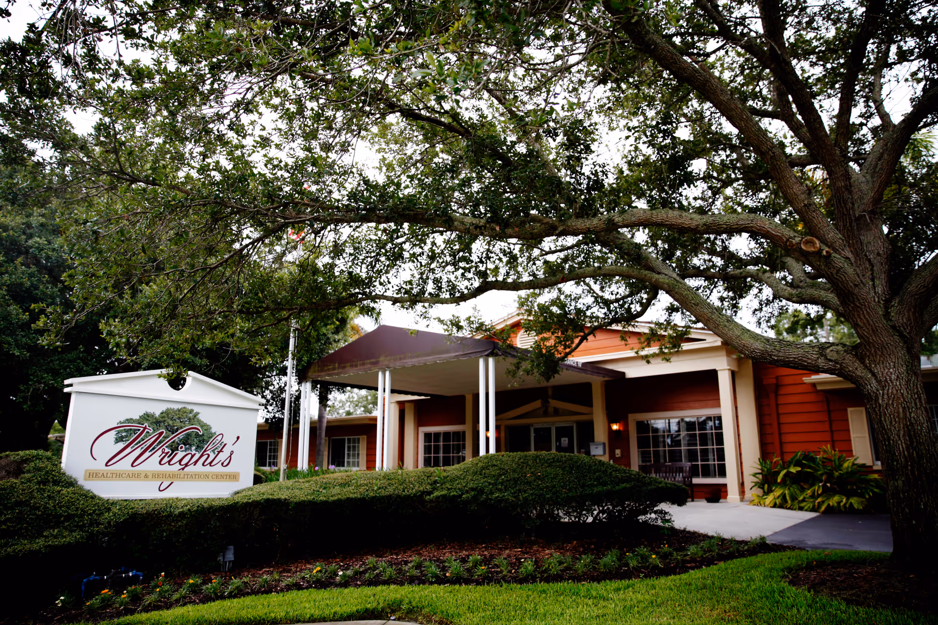 Front entrance of Wright's Healthcare & Rehabilitation Center with a large tree, canopy-covered porte-cochère, landscaped shrubs, and the facility sign.