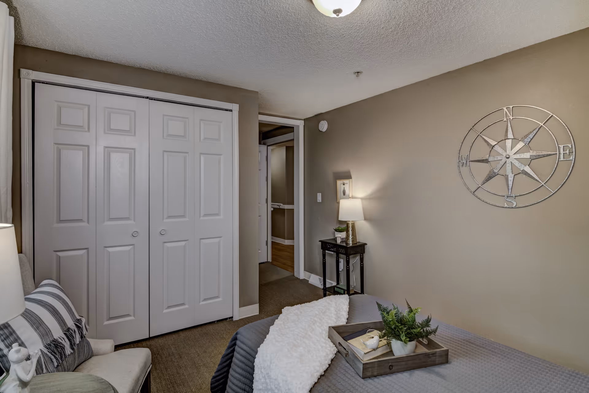 A cozy bedroom with a bed covered in gray bedding and a white throw blanket. A wooden tray with a small plant and decorative items sits on the bed. To the left, there is a cushioned chair with a striped pillow and a lamp beside it. The room has beige walls, a white double-door closet, and a small black side table with a lamp and a small plant. A decorative metal compass wall art is mounted on the wall above the side table.
