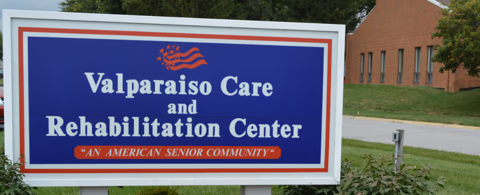 A large outdoor sign for Valparaiso Care and Rehabilitation Center with the tagline 'An American Senior Community' below the name. The sign has a blue background with white and red text, and an American flag graphic at the top. The sign is situated on a grassy area with a brick building and trees visible in the background.
