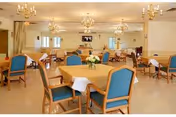 A spacious dining room with multiple tables covered with yellow tablecloths and white napkins. Each table is surrounded by wooden chairs with blue cushions. The room is well-lit with chandeliers hanging from the ceiling and has large windows along the walls. A television is mounted on the far wall.