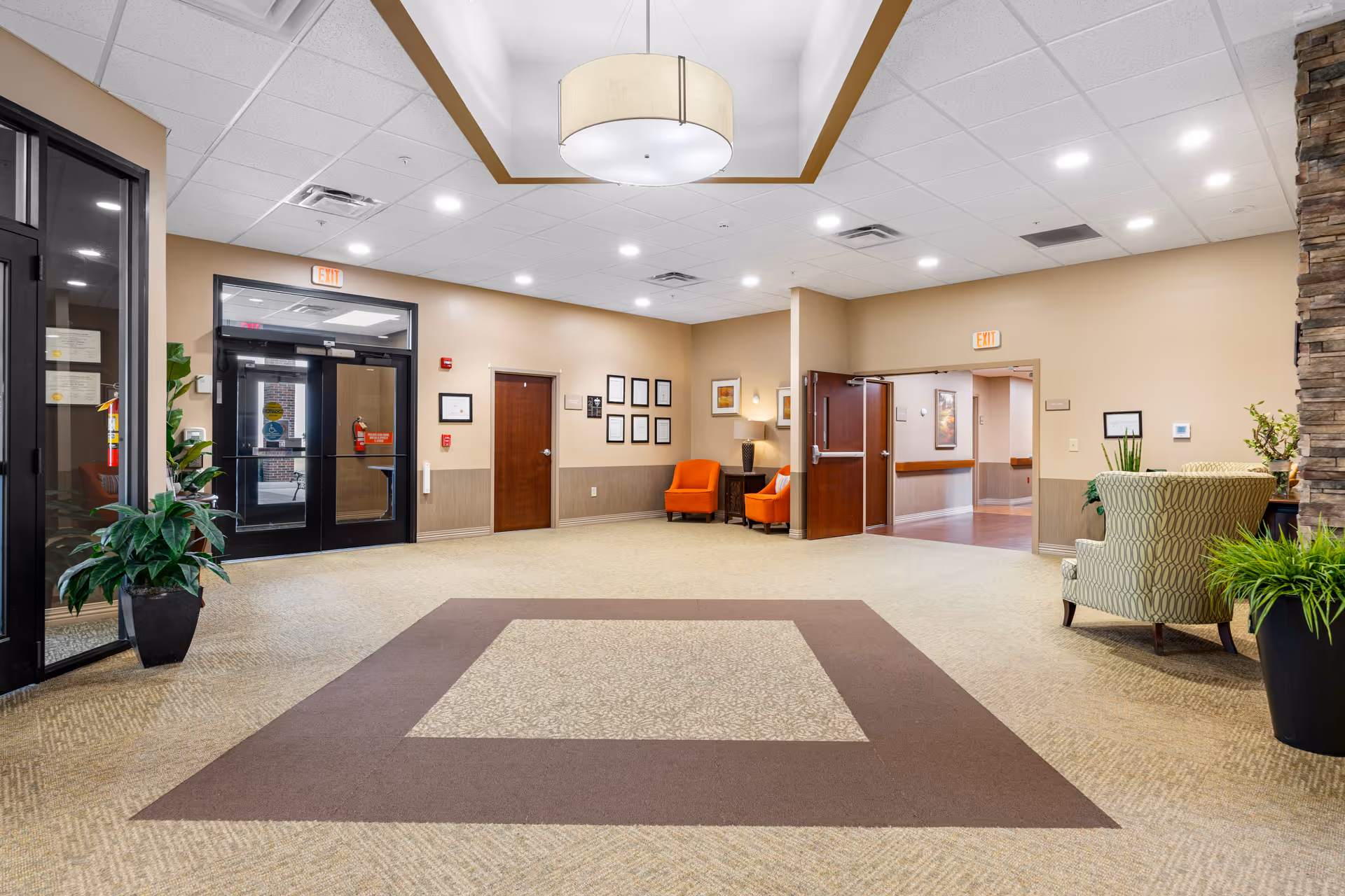 Spacious and well-lit lobby area of Vancrest of Payne featuring beige walls, a patterned carpet with a square design, a large ceiling light fixture, two orange chairs with a small table and lamp between them, a green patterned armchair, potted plants, and multiple doors including glass entrance doors and wooden interior doors.