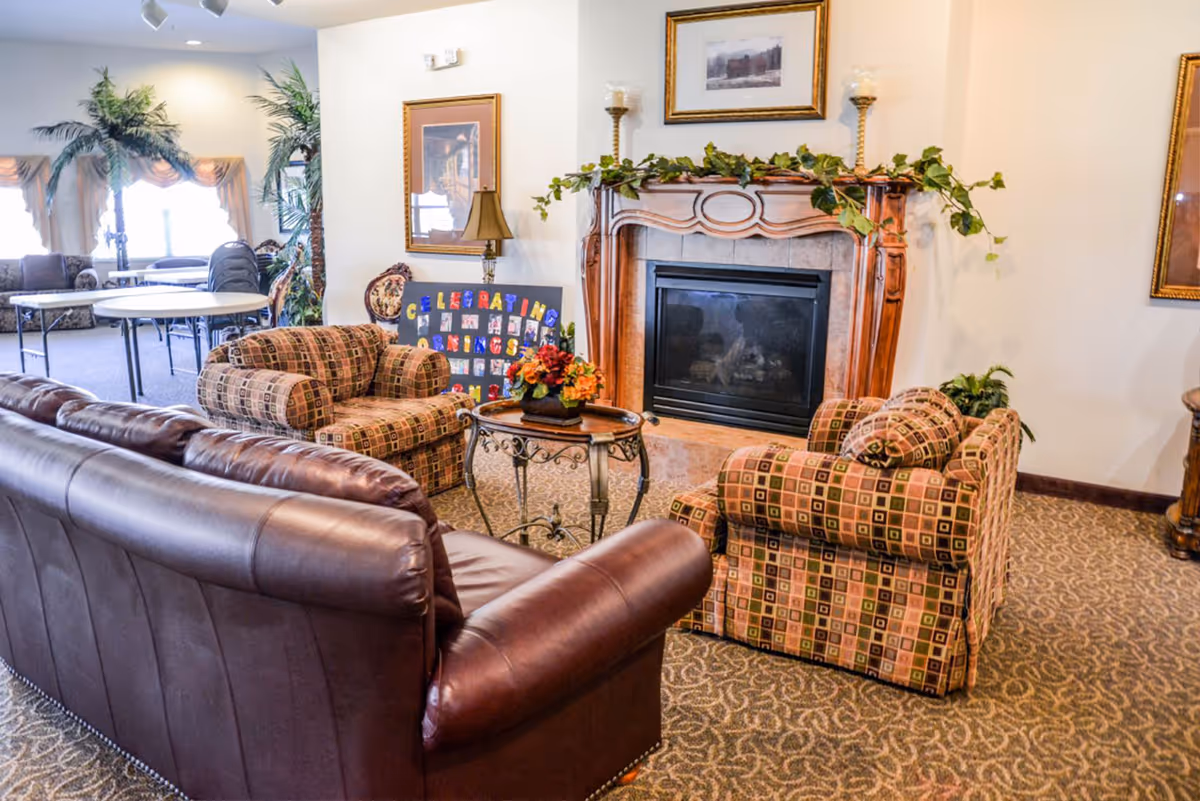 A cozy communal living room with a leather sofa, patterned armchairs, a decorative fireplace, and a small coffee table with flowers.