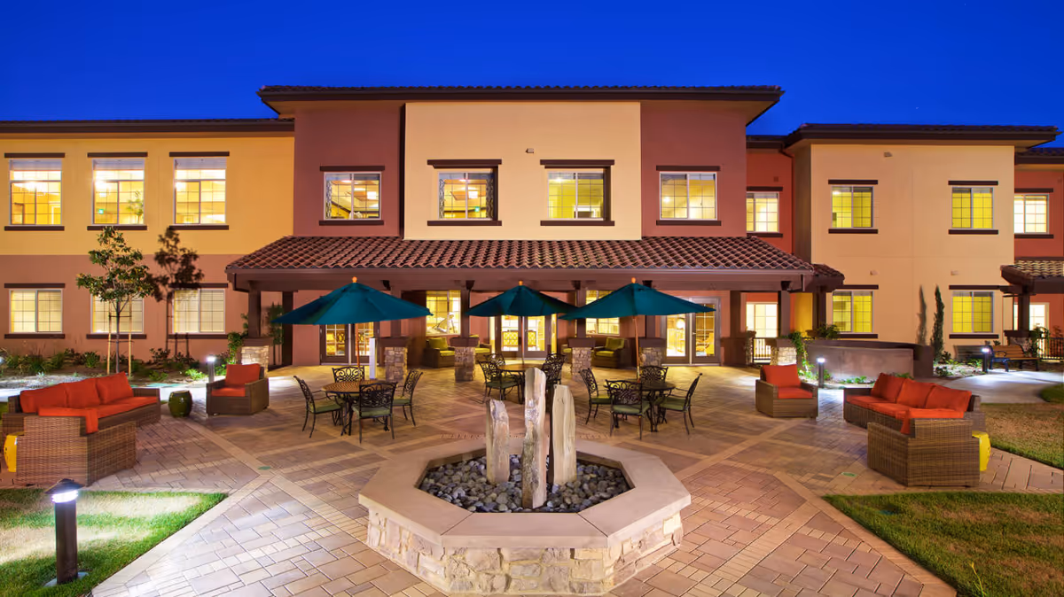 Well-lit courtyard with wicker seating, green umbrellas, and a central fountain in front of a two-story building.