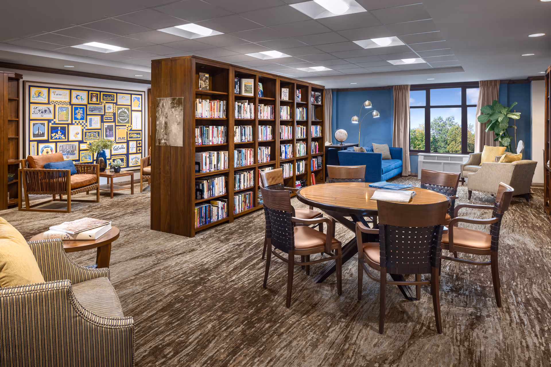 A bright senior living library/lounge with bookshelves, a round table and chairs, sofas and armchairs by a large window.