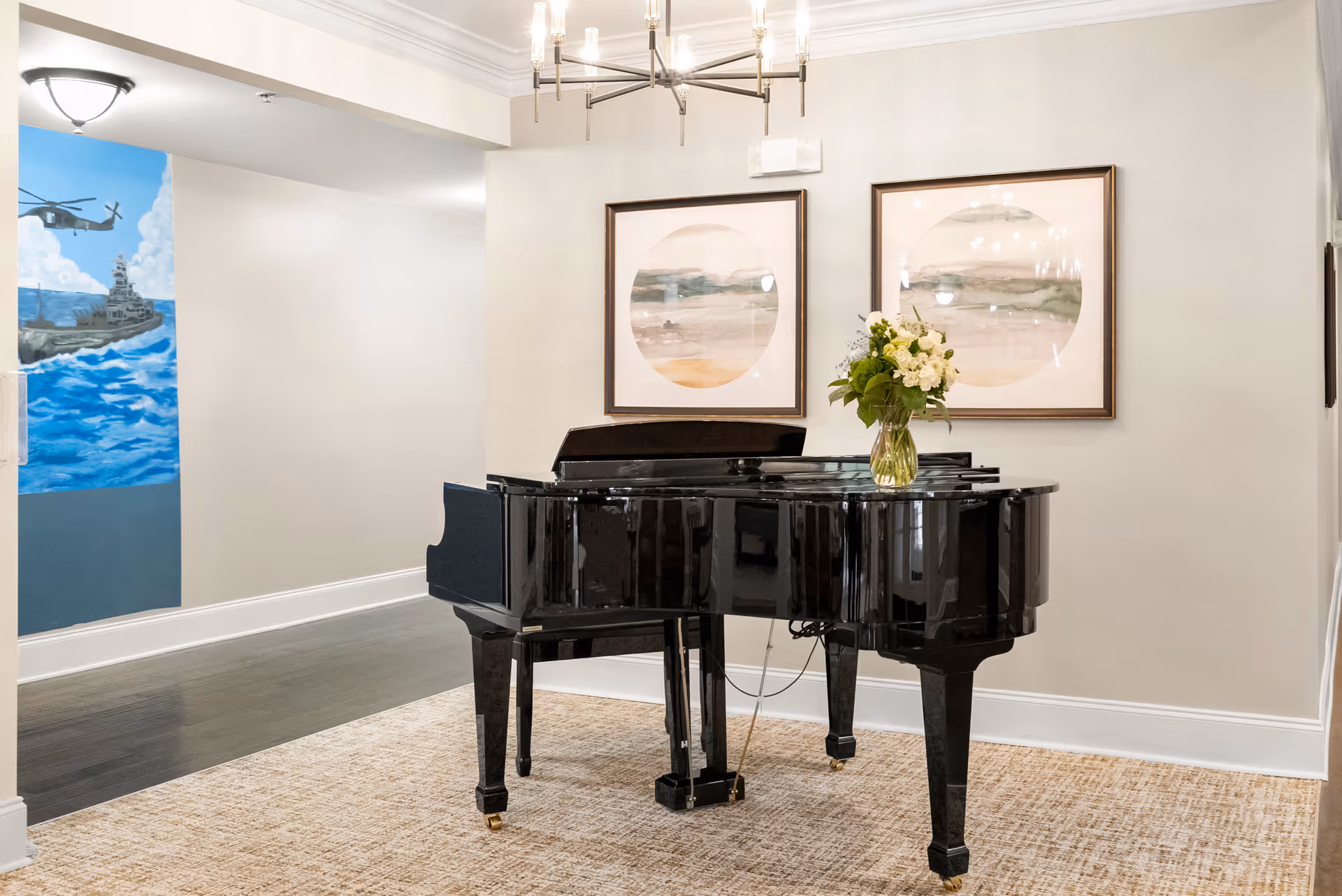 A black grand piano with a vase of white flowers on top is placed on a beige carpet in a well-lit room. Two framed abstract paintings hang on the light-colored wall behind the piano. A modern chandelier is mounted on the ceiling. In the background, there is a hallway with a mural depicting a helicopter and a ship over the ocean.