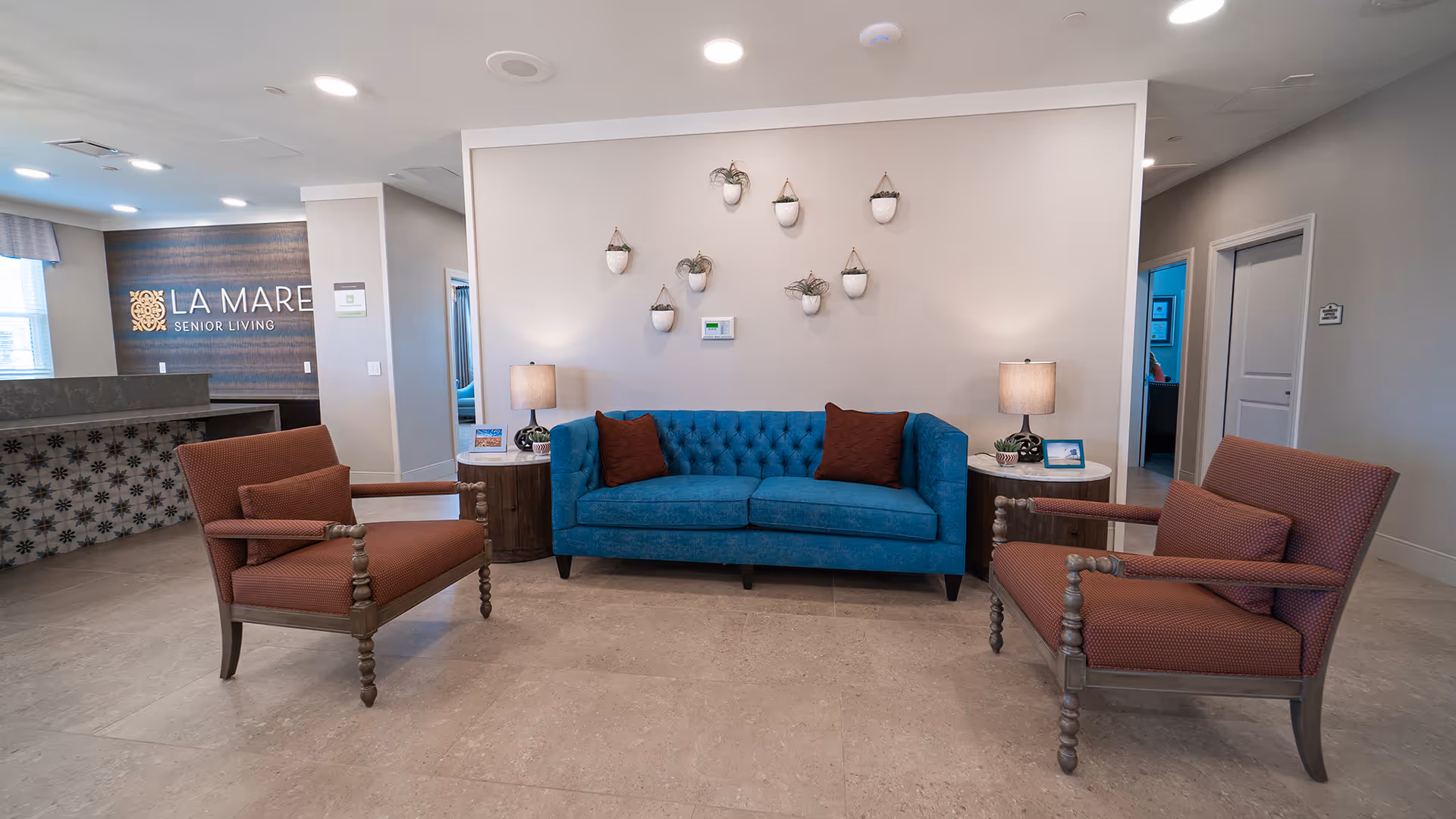 Lobby seating area with a teal tufted sofa, two upholstered armchairs, side tables with lamps, and wall planters.