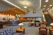 Spacious senior living facility lobby with a reception desk on the left, comfortable seating areas with armchairs and sofas, wooden tables with lamps, and a staircase leading to the upper floor. The area is well-lit with ceiling lights and has a modern, welcoming atmosphere.