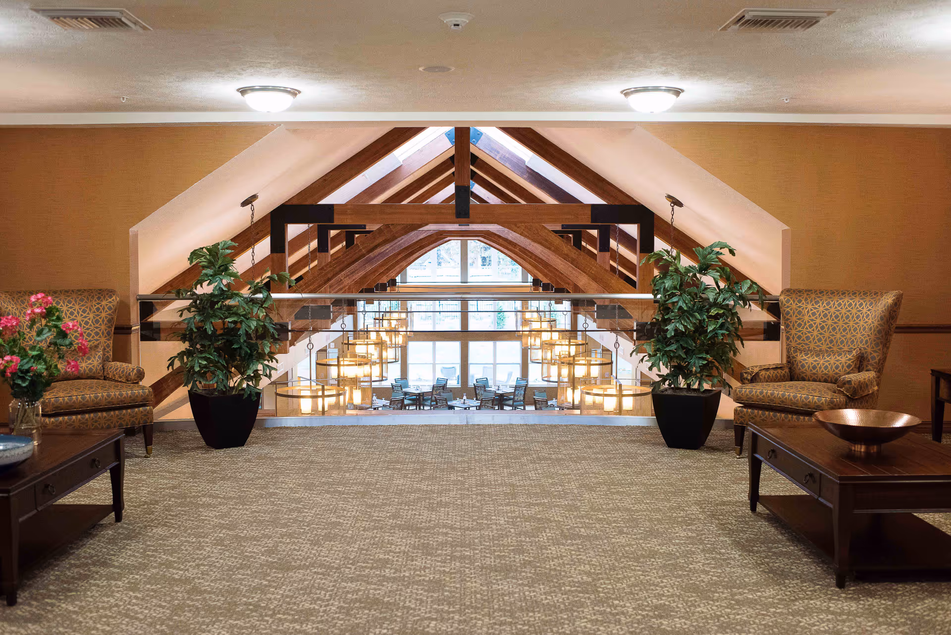 Upstairs lounge with armchairs, potted plants and coffee tables overlooking a light-filled atrium with exposed wooden beams and hanging chandeliers.