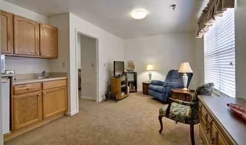 A cozy living area in an assisted living facility featuring a blue recliner, a floral upholstered chair, a wooden TV stand with a flat screen TV and a model ship, a side table with a lamp, and a window with blinds and a valance. To the left, there is a kitchenette with wooden cabinets and a sink.