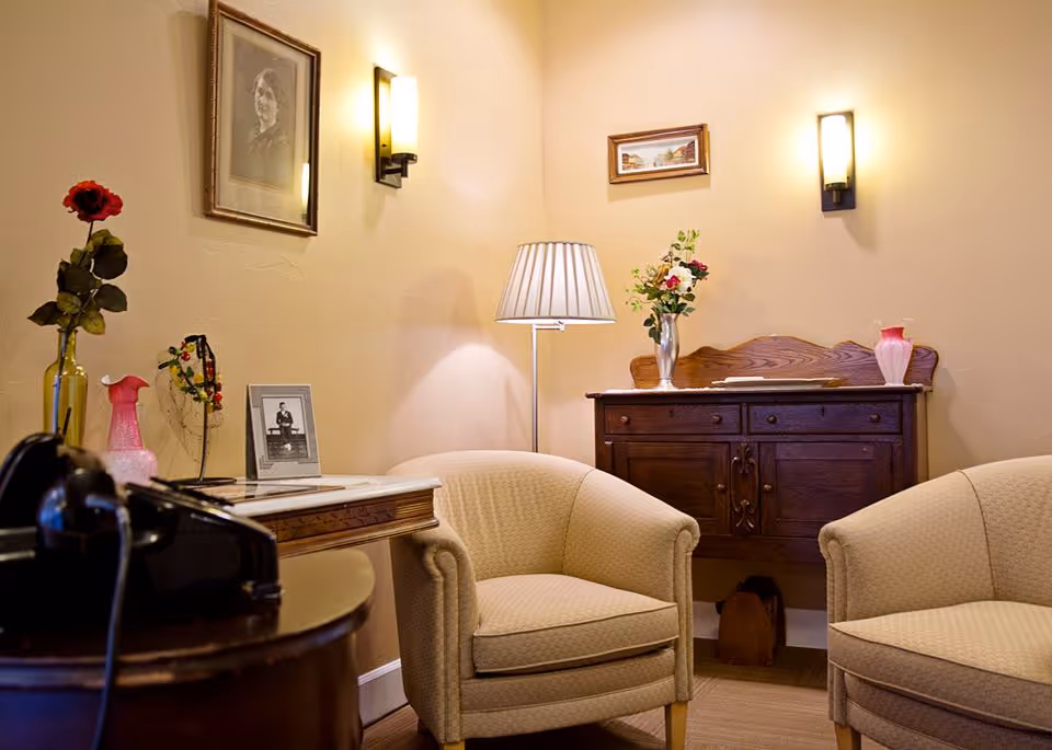 A cozy sitting area with two beige upholstered armchairs facing each other. Between them is a wooden side table with a vintage black rotary phone and a framed photograph. Behind one chair is a wooden cabinet with a vase of flowers and decorative items on top. The walls are painted a warm beige color and adorned with framed pictures and wall sconces emitting soft light. A floor lamp with a pleated shade stands in the corner.