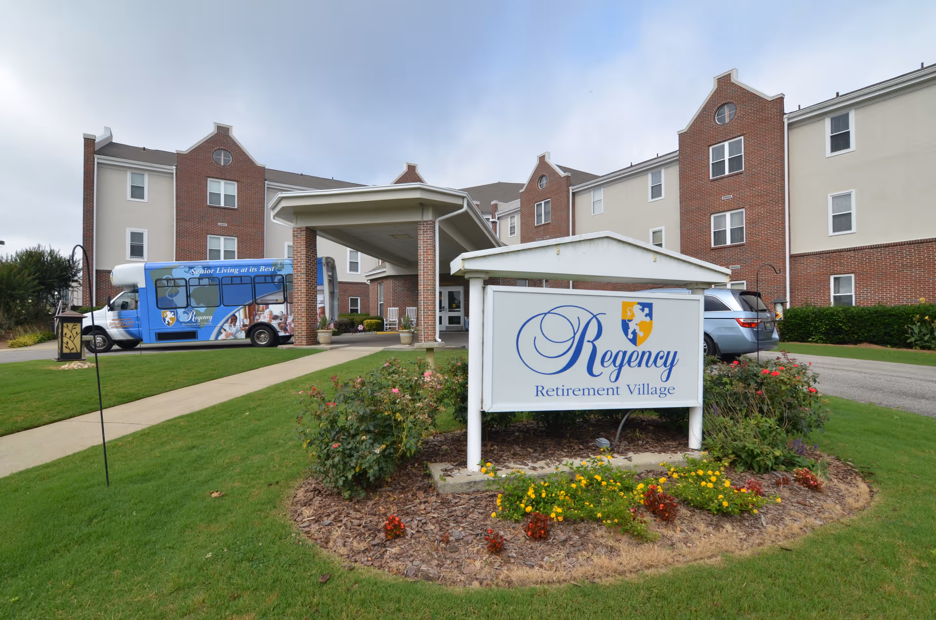 Front entrance of Regency Retirement Village with a sign, covered drop-off, shuttle bus, and landscaped lawn.