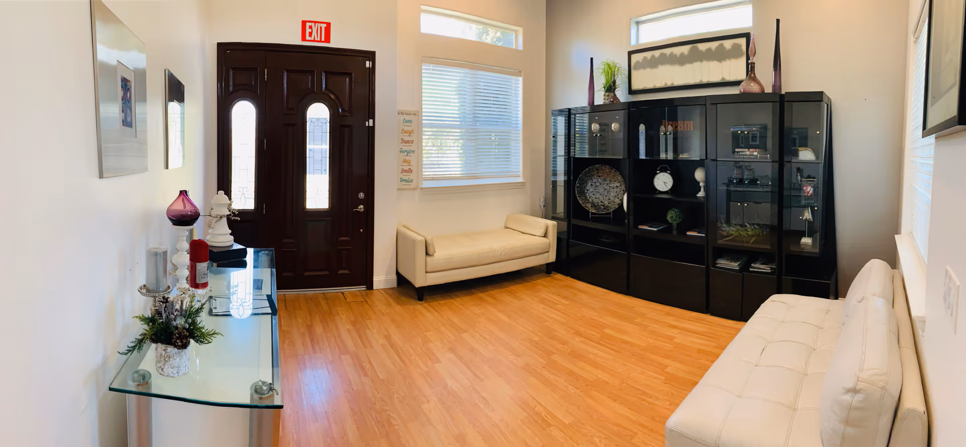 A bright and clean living room with light wood flooring, white walls, and modern furniture including two white cushioned benches and a glass console table with decorative items. A dark wooden front door with glass panels is visible, along with a large black display cabinet filled with decorative objects. Two windows with blinds allow natural light into the room, and an exit sign is mounted above the door.